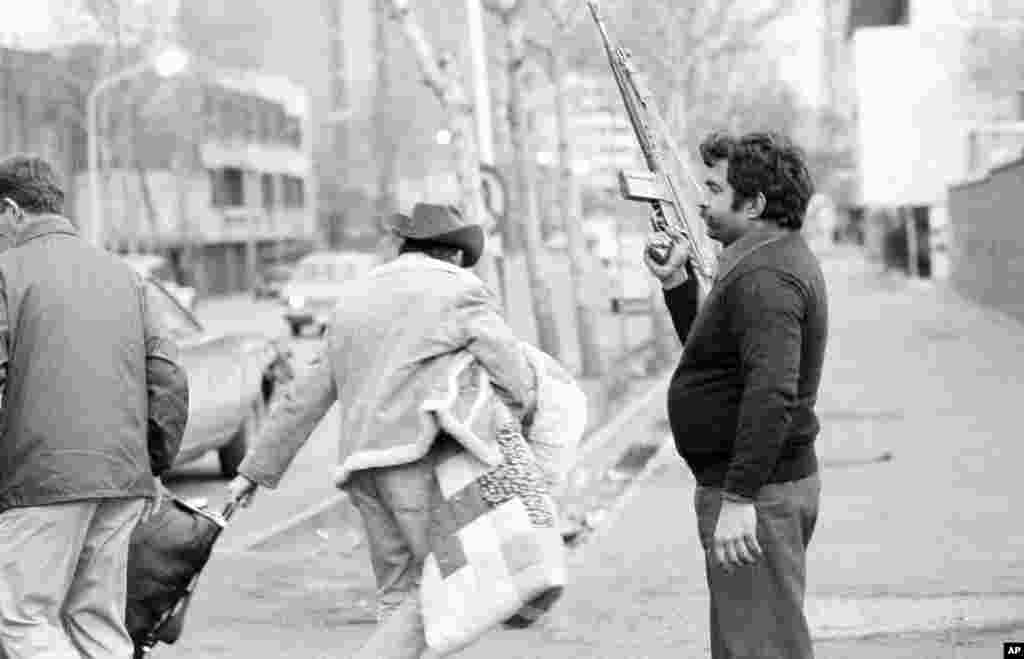 Američki državljani nose lične stvari prolazeći pored naoružanog Homenijevog lojaliste koji čuva prostor za evakuaciju u Teheranu 17. februara, 1979. godine. Stotine Amerikanaca napustile su Iran nakon prvog pokušaja zauzimanja ambasade.