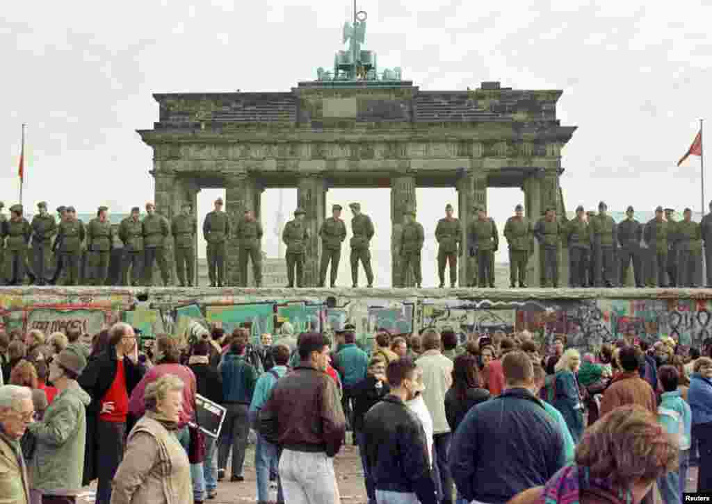 Graničari istočnog Berlina stoje na vrhu Berlinskog zida ispred Brandenburških vrata 11. novembra 1989. Dva dana ranije, 9. novembra, zid su probile hiljade Nijemaca nakon što je dužnosnik istočne Njemačke rekao da su ograničenja putovanja između istoka i zapada Njemačke trebala biti okončana &quot;bez odlaganja&quot;.